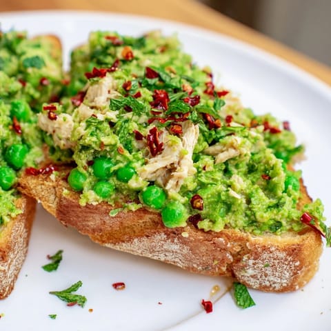 A close-up view of the Smashed Pea & Chicken Toast, showcasing its chunky texture and colorful garnishes on a rustic wooden board.